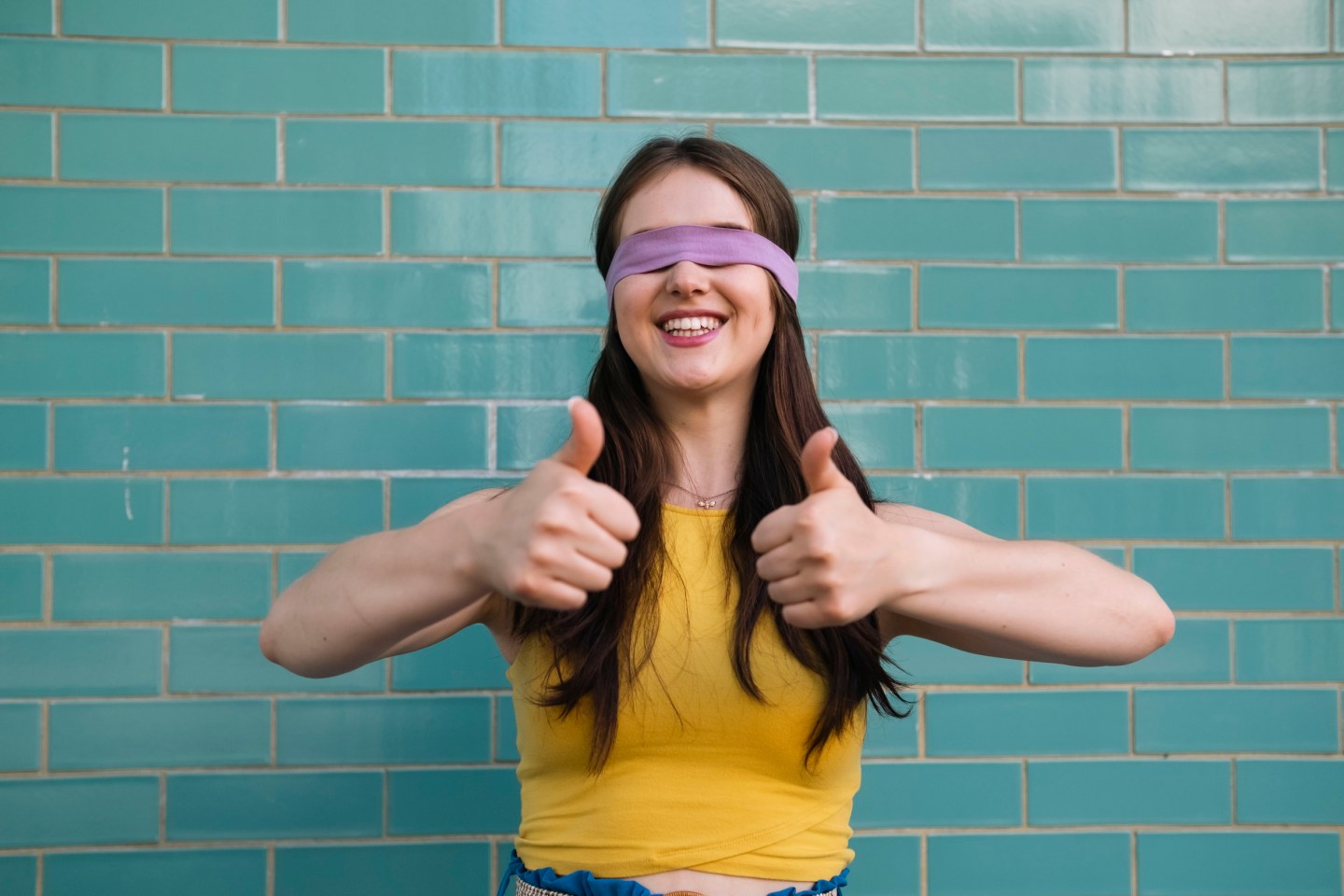 Smiling woman with blindfold showing thumbs up gesture in front of teal wall