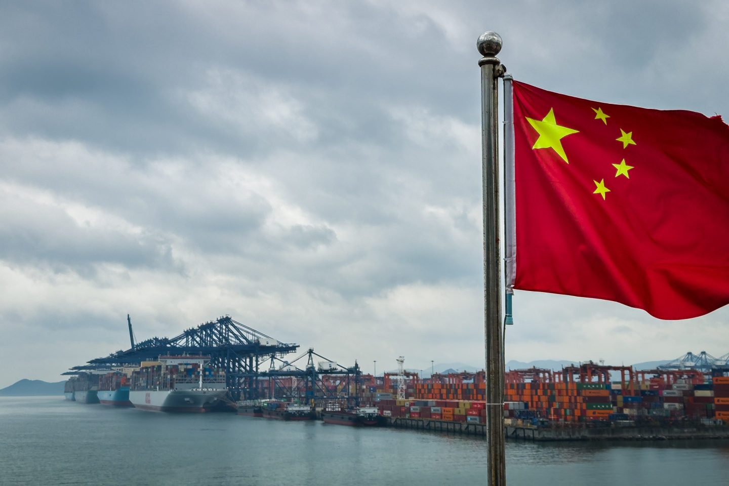 A Chinese national flag is seen in the foreground with container ships, cranes, and stacked shipping containers at the Yantian International Container Terminal under cloudy skies, on April 12, 2025 in Shenzhen, China.