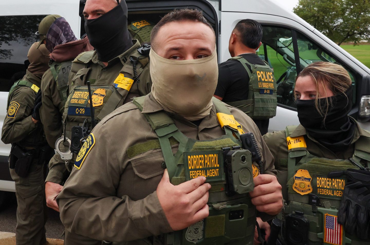 A man wearing a "border patrol" best has his face covered with a mask and is looking forward.
